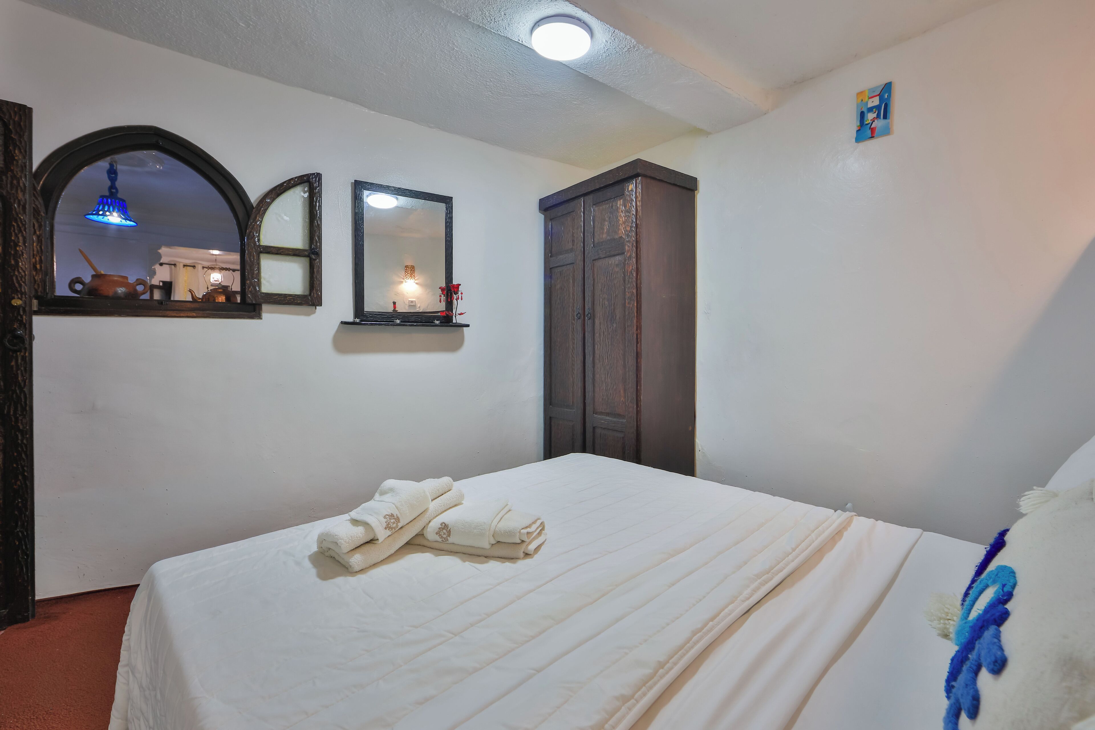Ein ordentlich bezogenes Bett mit Handtüchern, ein Holzschrank, ein Spiegel und eine kleine Ablage mit einer Lampe.