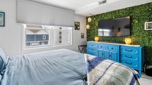 Quarto com cama, cómoda azul, parede verde, televisão e janela com persianas.