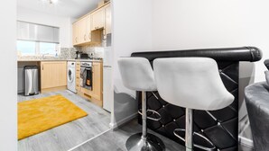 A compact kitchen with modern appliances, a washing machine, and a coffee machine. There are two bar stools with cushioned seats and metal legs. A yellow rug is on the floor, and the kitchen has a window with blinds. - Stunning 2-bed Apartment in Tipton Sleeps 3 (Tipton)