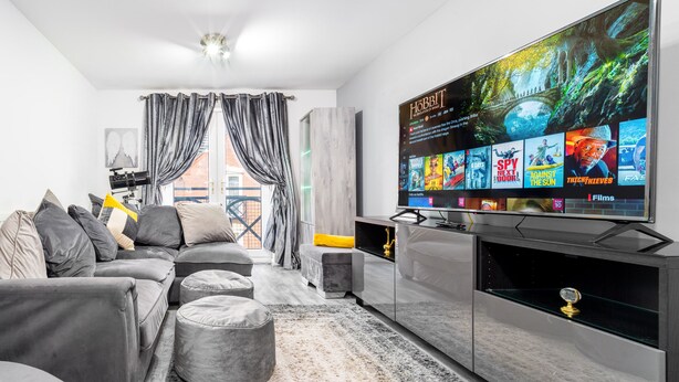 A modern living room with a large flat-screen TV displaying movie options, grey sofas, ottomans, and a fireplace. - Stunning 2-bed Apartment in Tipton Sleeps 3 (Tipton)