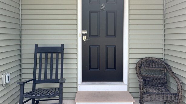 A black door with the number 2, a wooden bench, and a wicker chair outside a house with light green siding. - Three Schooners Landing - Middle Condo (Sodus Point)