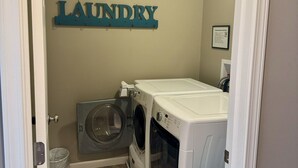 A laundry room with a wall-mounted sign reading "LAUNDRY", two washing machines, a dryer, and a blue laundry basket. - Three Schooners Landing - Middle Condo (Sodus Point)