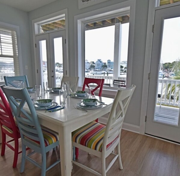 A dining area with a white table set for four, colorful chairs, and large windows offering a view of the marina. - Luxury canal front 3 bedroom condo with boat dock and central location (Carolina Beach)