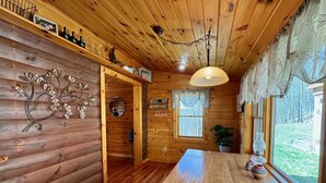Ein Holzblockhaus im Inneren mit einem Esstisch, Holzwänden, einer Deckenleuchte, einem Fenster mit Blick ins Freie und einem Wandbild.
