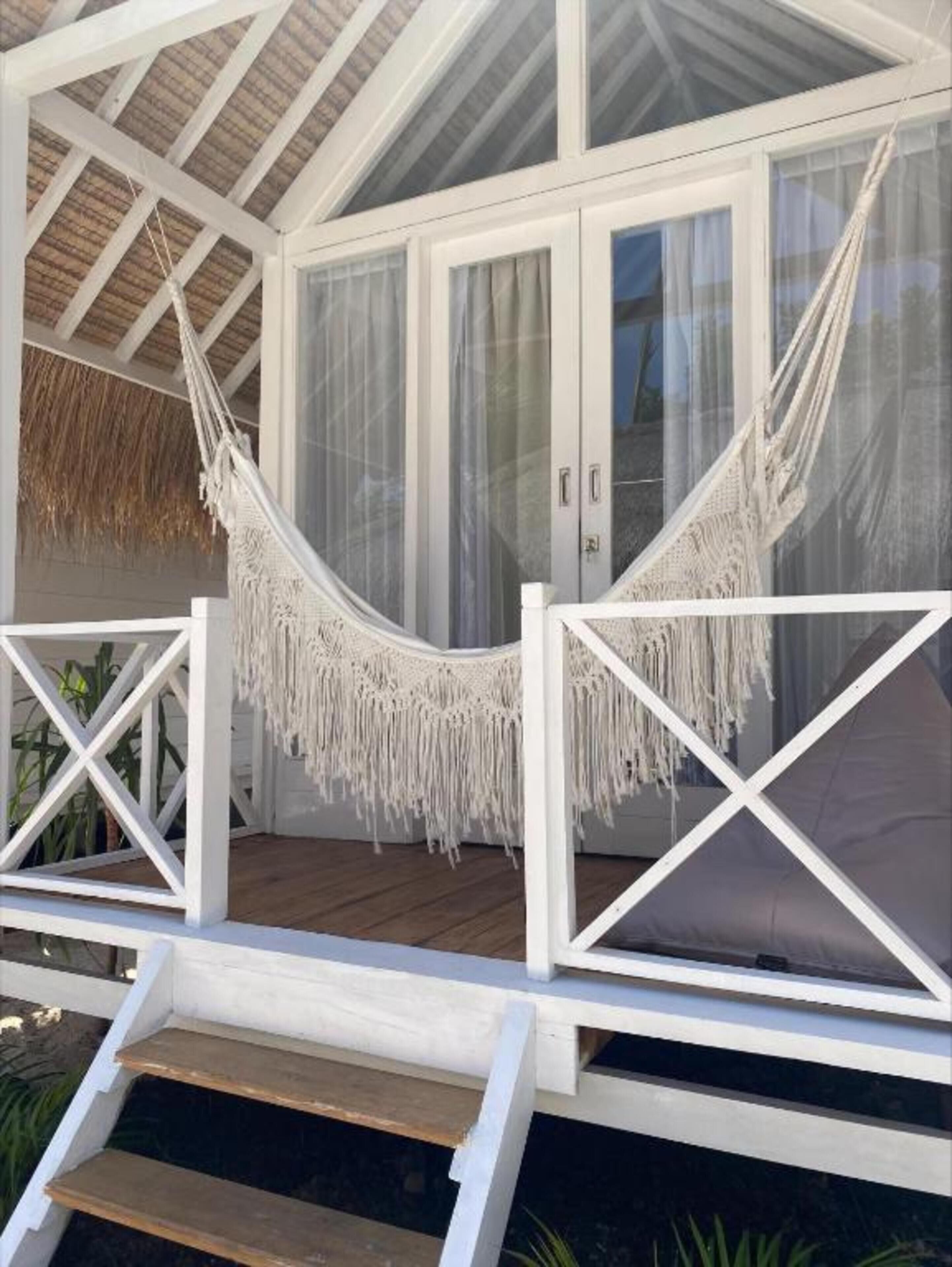 A covered outdoor area with a white hammock, wooden deck, and glass doors leading to a room.