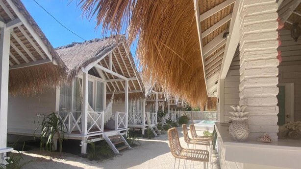 Un complejo turístico frente a la playa con estructuras de techo de paja, mobiliario exterior y un patio arenoso.