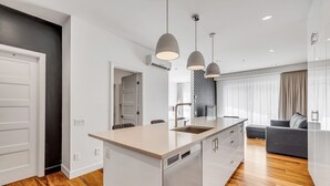 A modern kitchen with a central island, pendant lighting, and a view to a living area with a sofa and curtains. - Initial Ilot Downtown Québec (Québec City)