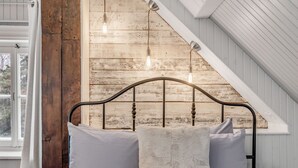 A bedroom with a sloped ceiling, wooden wall paneling, a metal bed frame, and three hanging light fixtures. - Initial | La petite école | Île d'Orléans (Saint-Laurent-de-l'Île-d'Orléans)