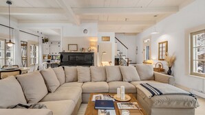 A spacious living room with a large beige sectional sofa, a wooden coffee table with magazines and candles, and a piano in the background. - Initial | La petite école | Île d'Orléans (Saint-Laurent-de-l'Île-d'Orléans)