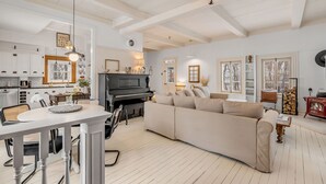 A spacious living room with a piano, a beige sofa, and a dining area with a white table and chairs. - Initial | La petite école | Île d'Orléans (Saint-Laurent-de-l'Île-d'Orléans)