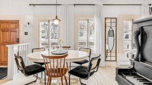 A dining area with a round table, black chairs, and a piano. - Initial | La petite école | Île d'Orléans (Saint-Laurent-de-l'Île-d'Orléans)