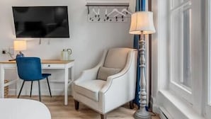 Hotellihuone, jossa on valkoinen nojatuoli, työpöytä lampun kanssa, televisio ja ikkuna, jossa on verhot.