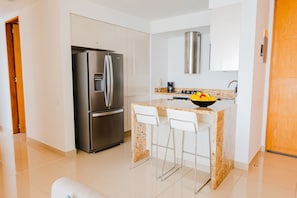 A modern kitchen with a stainless steel refrigerator, a granite countertop, white bar stools, and a built-in oven.