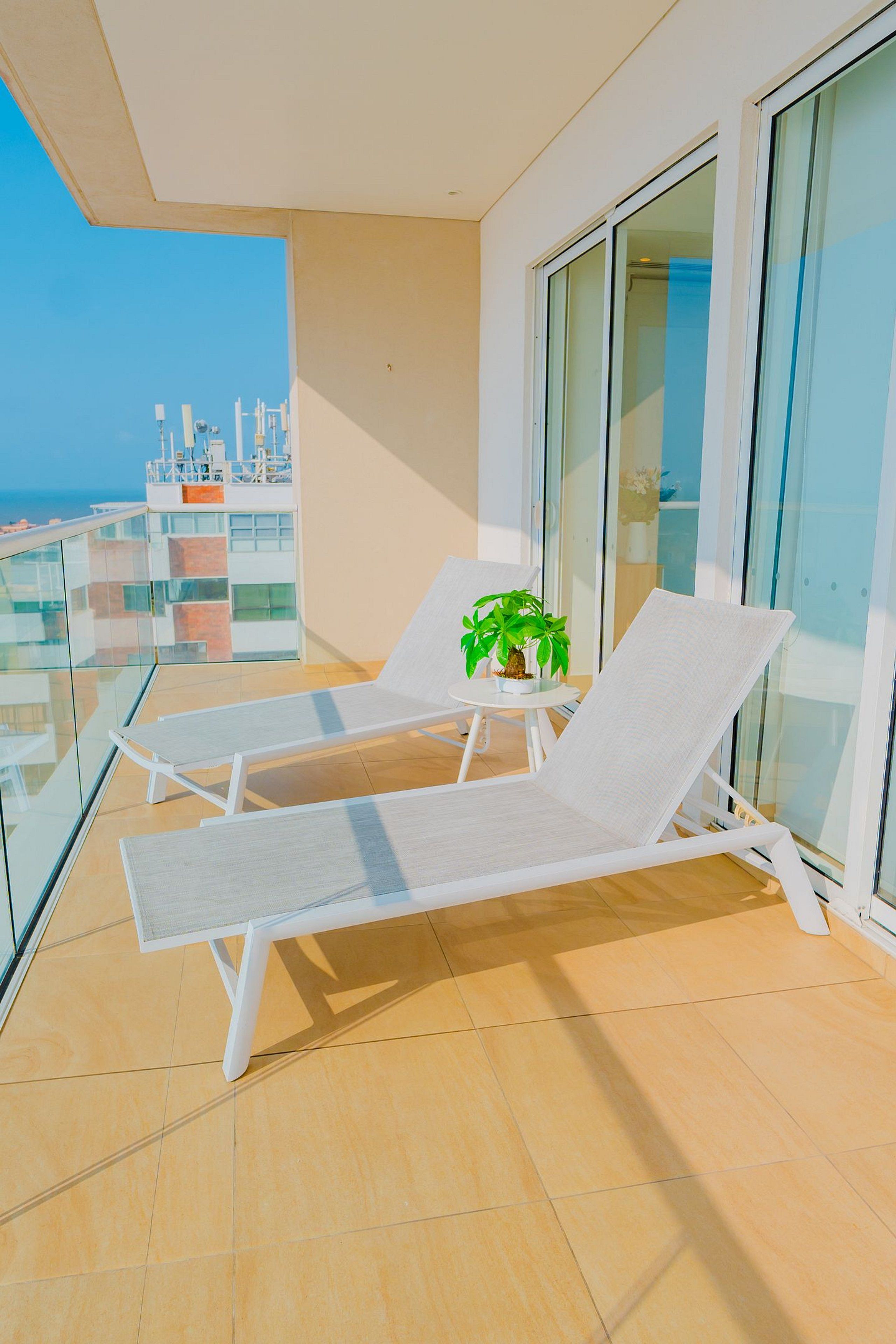A balcony with two white lounge chairs, a small table with a potted plant, and a glass railing.