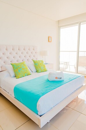 Habitación de hotel con una cama grande, cabecera blanca y vistas al exterior.