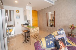 Cocina compacta con lavamanos, estufa y refrigerador. Hay un taburete pequeño y un espejo decorativo en la pared.
