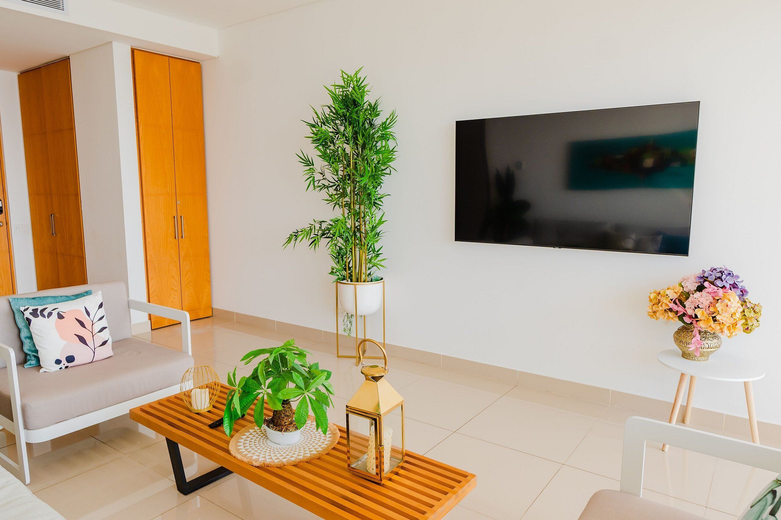 Sala de estar moderna com sofá, mesa de centro com objetos decorativos, planta em vaso, TV fixada na parede e pequena mesa com um vaso de flores.