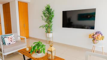 Sala de estar moderna com sofá, mesa de centro com objetos decorativos, planta em vaso, TV fixada na parede e pequena mesa com um vaso de flores.