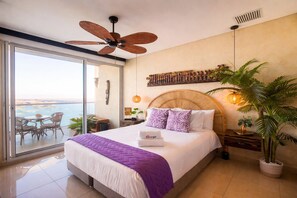 Ein modernes Schlafzimmer mit einem großen Bett, einem Deckenventilator, Meerblick und einem Balkon mit Sitzgelegenheiten.