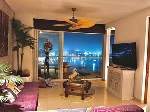 Una sala de estar con un televisor grande, un ventilador de techo, un sofá, una mesa de centro y vistas a un paisaje urbano a través de un vidrio.