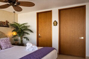 Un dormitorio con cama, ventilador de techo, repisa de pared con objetos decorativos, planta en maceta y dos puertas de madera.