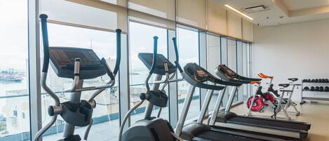 Ett gym med löpband, crosstrainers och en motionscykel, stora fönster och utsikt över en stad.