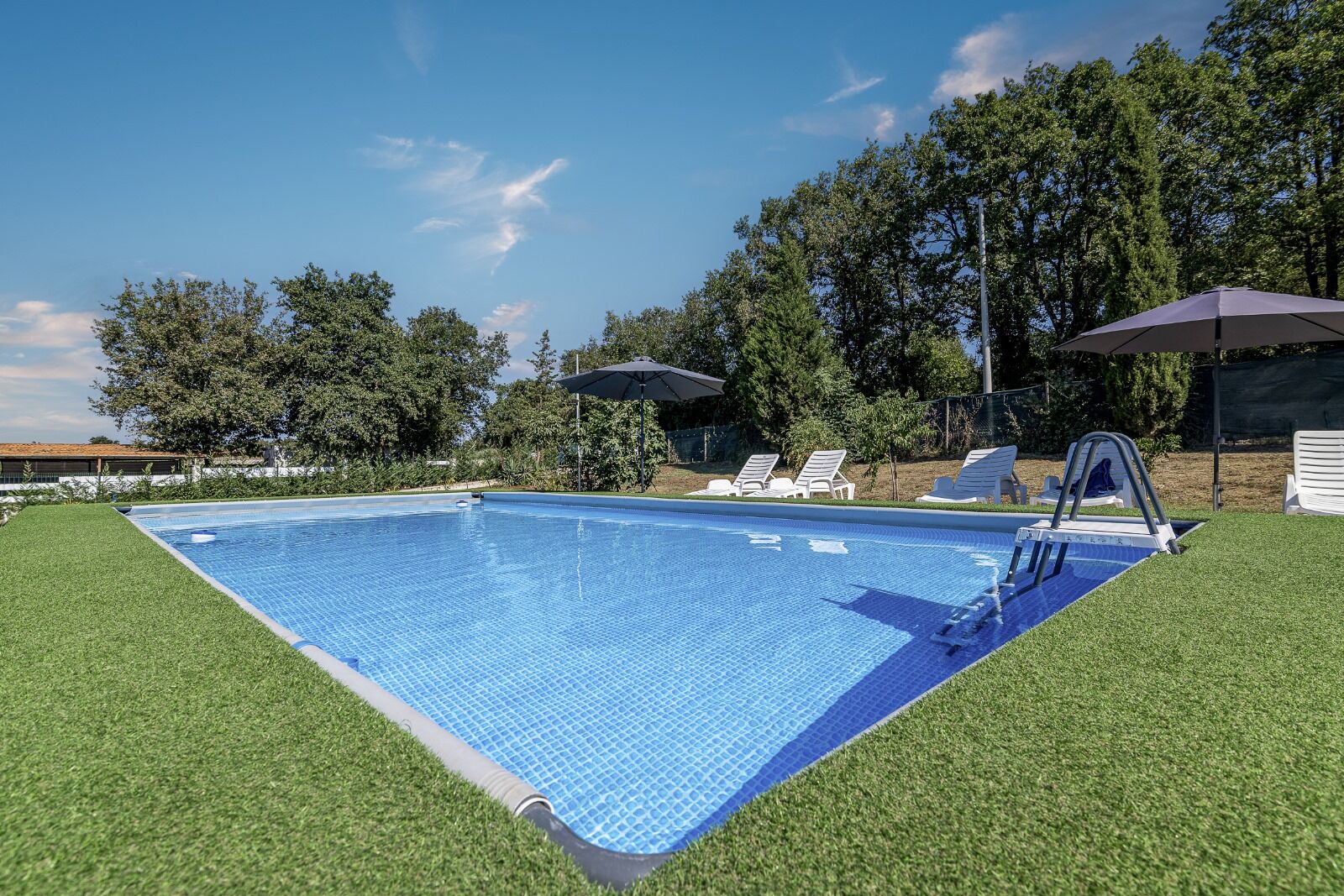Une piscine rectangulaire aux parois recouvertes de carreaux bleus, entourée de gazon synthétique. On aperçoit des chaises longues et des parasols à l’arrière-plan.