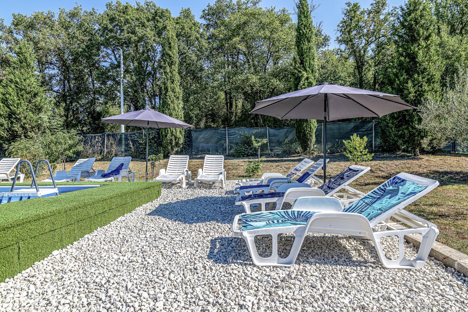 Zona de piscina con sillas reclinables blancas y sombrillas, rodeada de árboles y una valla delimitando la piscina.