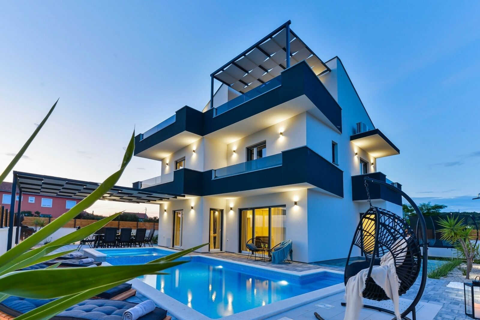 Una casa moderna de dos pisos con piscina, mobiliario exterior y una pérgola.