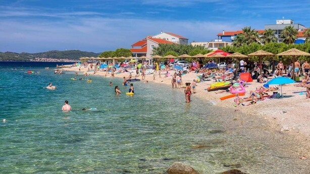 A beach with clear shallow waters, umbrellas, and beachgoers. - Holiday Home in Bibinje with Seaview, Balcony, Air condition, WIFI (5201-1) (Bibinje)