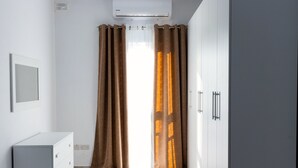 A modern room with a white wardrobe, a small white cabinet, and a large window with brown curtains. - Seaside Serenity - Luxe Penthouse - near the beach (Il-Mellieha)