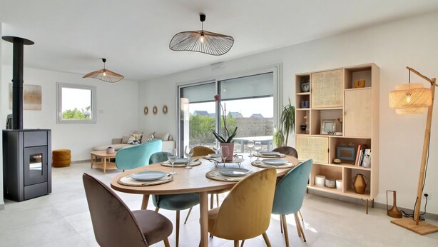 A modern dining area with a round table set for four, surrounded by chairs. There is a black stove in the corner, a large window providing a view of the outdoors, and a wooden bookshelf with various items on the right side. - Maison 6 Personnes, SPA (Penmarc'h)
