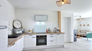 A modern kitchen with white cabinets, a built-in oven, a microwave, and a sink. There is a wooden countertop with a bowl of fruit and a bottle of orange juice. A clock and a small potted plant are on the wall. - Maison 6 Personnes, SPA (Penmarc'h)