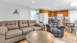 Ein modernes Wohnzimmer mit Sofa und Sesseln, ein Essbereich mit Holzsäulen und eine Küche mit Kühlschrank und Mikrowelle.
