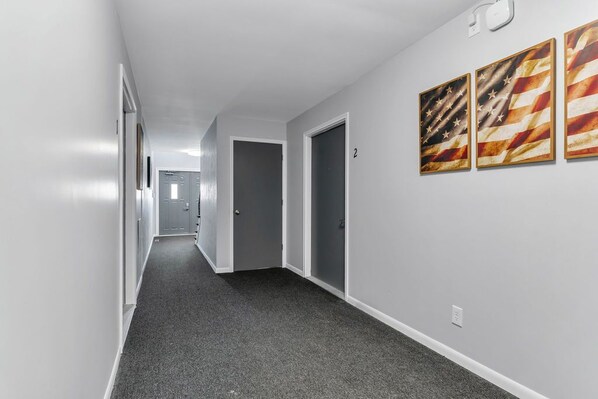 A hallway with grey carpet, white walls, and three framed American flag pictures on the right wall.