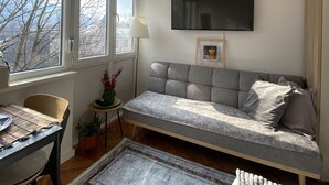 Een woonkamer met een bank, een aan de muur bevestigde televisie, een eettafel met stoelen en een raam met uitzicht op bomen en gebouwen.