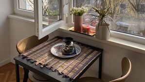 Een eethoek met een tafel gedekt voor twee personen, een raam met uitzicht op kale bomen en gebouwen, en een klein schap met een potplant en een kaars.