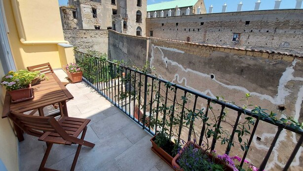 A balcony with a table and chairs, potted plants, and a view of old stone buildings. - Carmine's Balcony (Napoli)