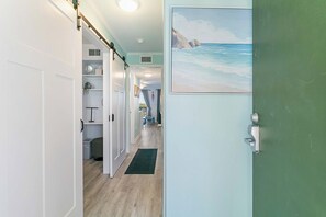 A hallway with a view of a beach painting, a door with a handle, and a shelf with items.