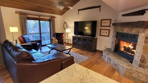 A living room with a stone fireplace, leather sofa, television, and a view of the outdoors through a large window. - Sapphire Mountain View Escape: Resort Amenities, Family & Pet Friendly! (Sapphire)