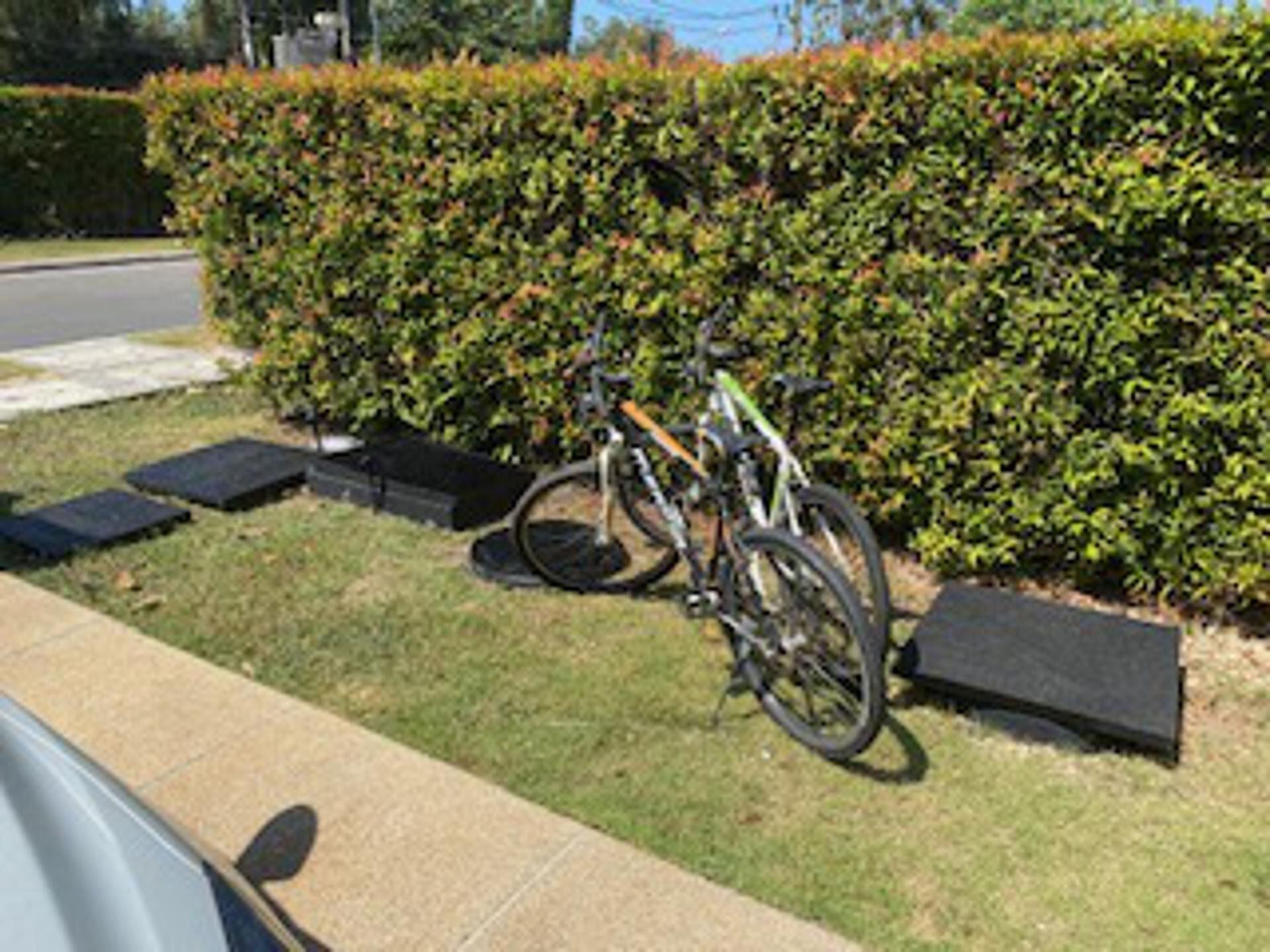 A bicycle parked next to a hedge, with a sidewalk and a road in the background.