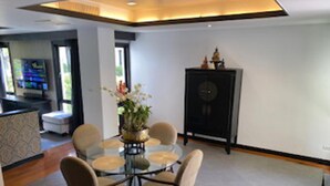 A dining area with a glass table surrounded by chairs, a cabinet with a decorative item on top, and a television in the background. - Poolside Luxury Laguna 2-bed Villa in Cherngtalay (Choeng Thale)