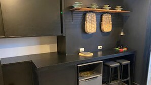 A modern kitchen with dark cabinetry, a countertop, and a small sink. There are two stools and a shelf with dishes. - Poolside Luxury Laguna 2-bed Villa in Cherngtalay (Choeng Thale)