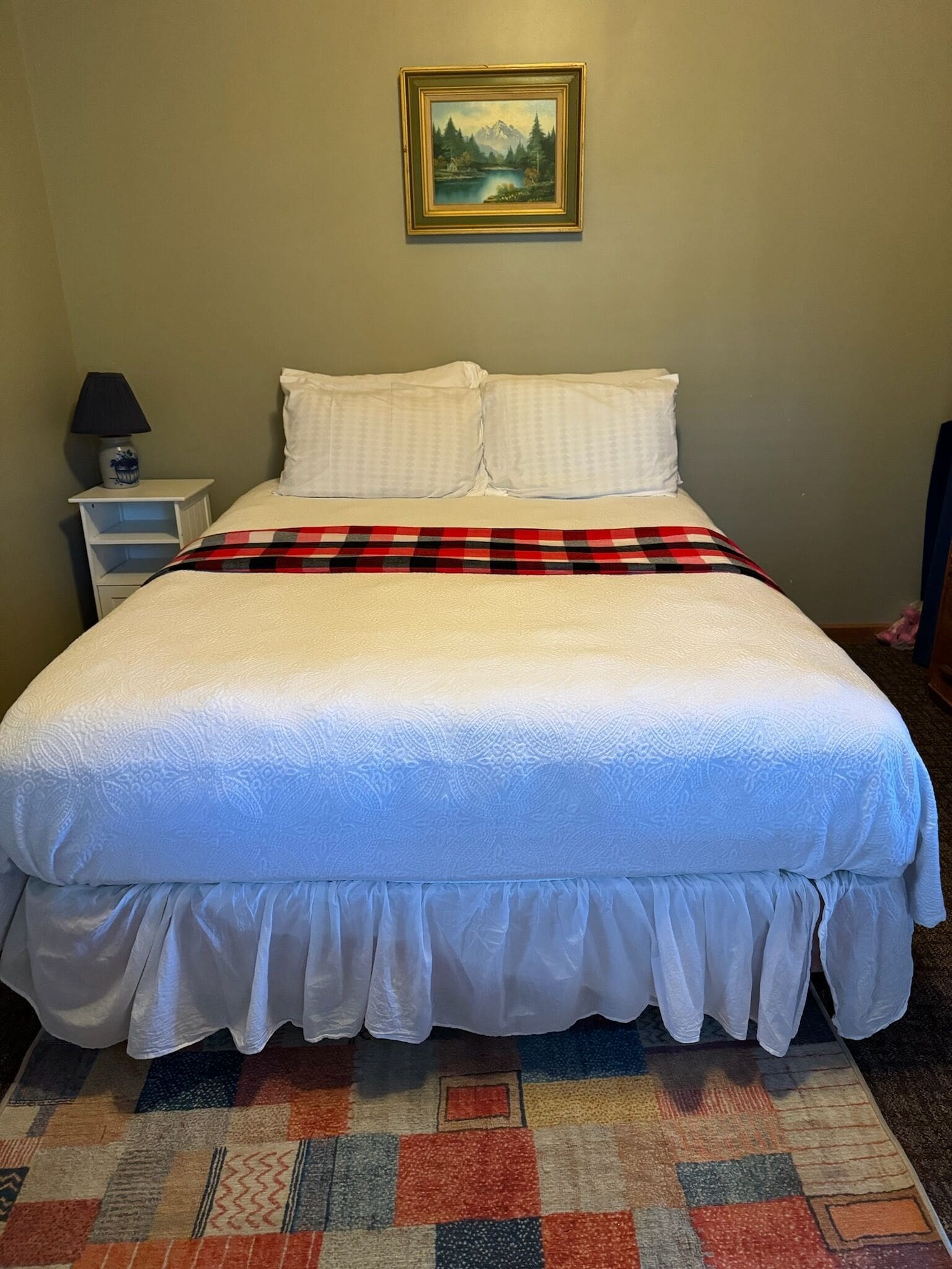A neatly made bed with white bedding and a plaid blanket, a small bedside table with a lamp, and a framed picture on the wall.