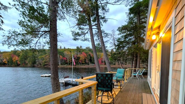 Terraza de madera con mobiliario exterior, con vistas a un lago con barcos y árboles.
