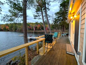 Une terrasse en bois avec des sièges extérieurs, donnant sur un lac où l’on peut voir des bateaux et des arbres.