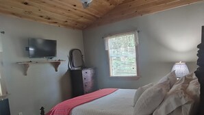 Un dormitorio con techo de madera, una cama con ropa de cama blanca y roja, una mesita de noche, una lámpara, un televisor en la pared y una ventana con persianas.