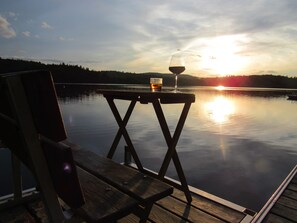Una terraza de madera con una copa de vino y una cerveza sobre una mesa, con vistas a un lago tranquilo y un atardecer de fondo.