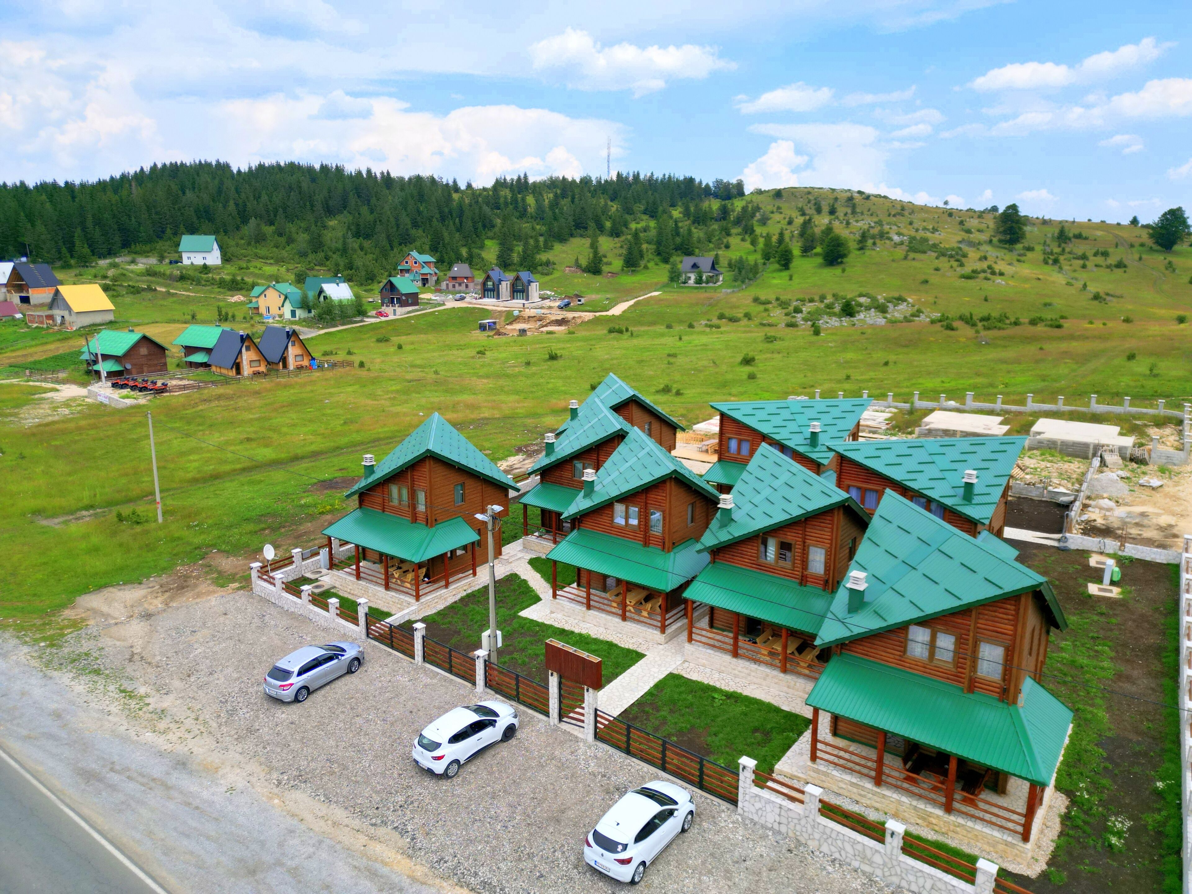 A row of wooden cabins with green roofs, arranged in a row on a grassy field with a forested hill in the background.
