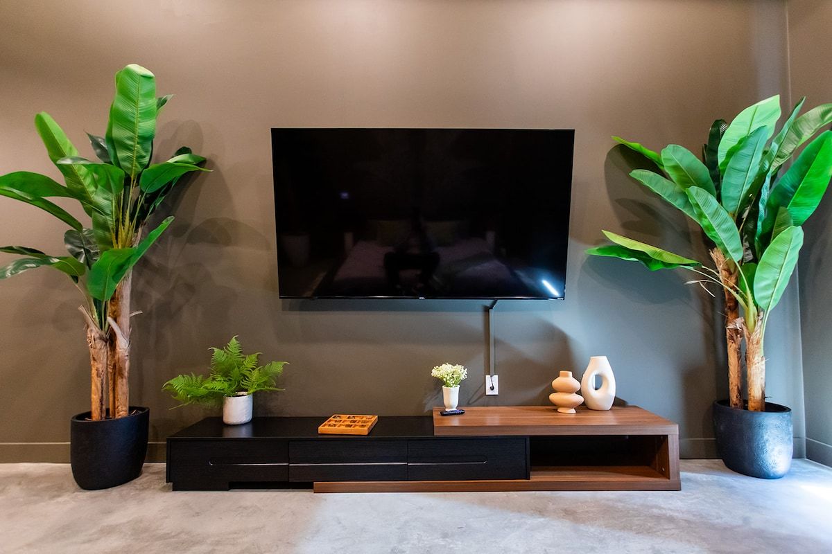 A modern living room with a flat-screen TV mounted on a wall, a black entertainment unit below, and two large potted plants on either side.